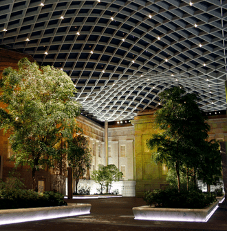The Kogod Courtyard at night with uplit marble bench walls and trees beneath the geometric glass ceiling