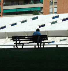 David Malda sits on a bench in shadow. Farther away, there is a brightly lit white angular wall.