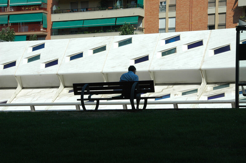 David Malda sits on a bench in shadow. Farther away, there is a brightly lit white angular wall.