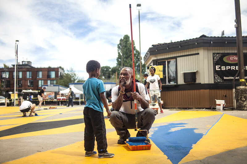 An older man holding a paint roller on a long handle squats down to take a break from painting blue triangles on the pavement to talk with a young boy