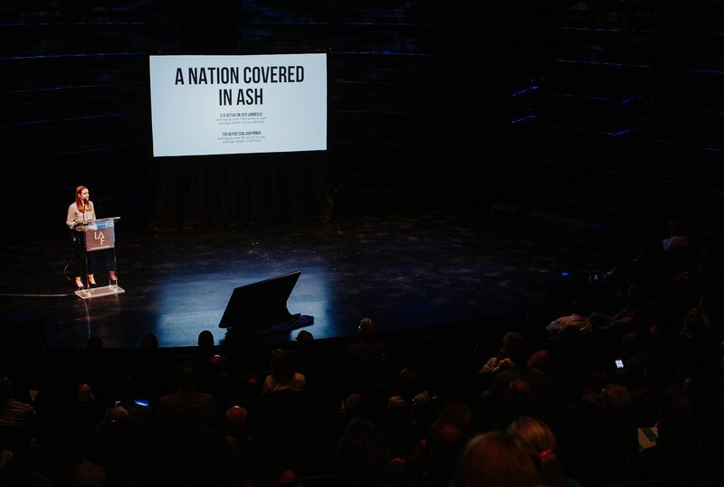 Lauren Delbridge presents to a packed audience. Behind her is a slide with the text "A NATION COVERED IN ASH".