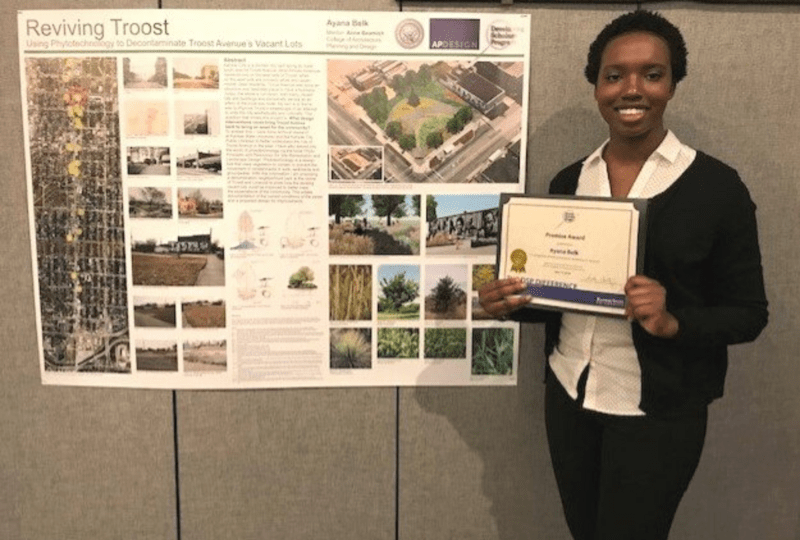 Ayana Belk holding a certificate award in front of a poster for her Reviving Troost work