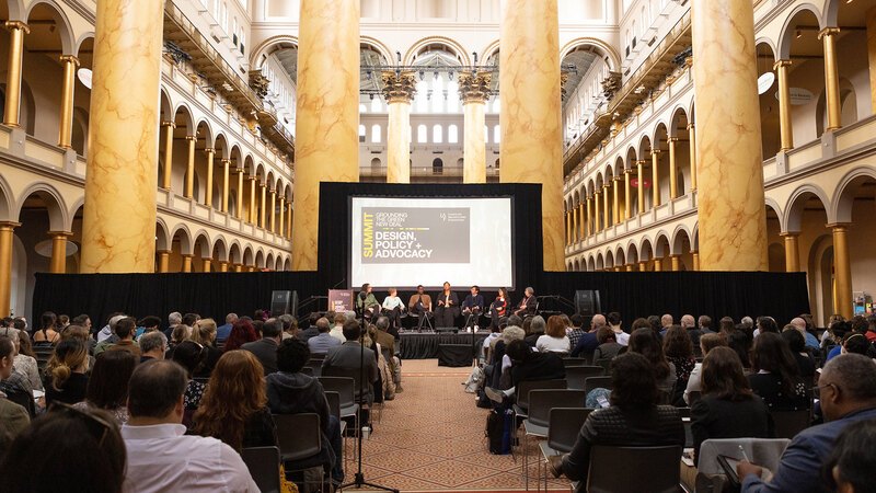 The stage, screen, and panelists in front of a seated audience amidst the columns and arches of the National Building Museum's Great Hall