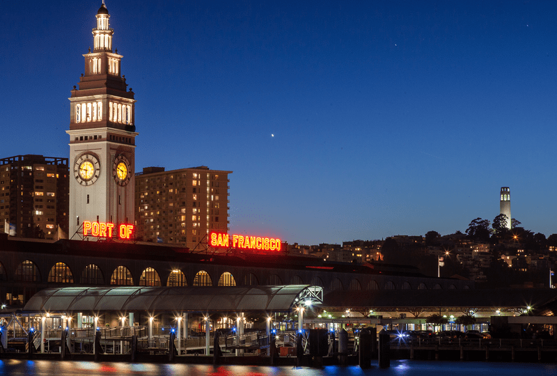 The illuminated clock tower, arches, and words "Port of San Francisco" along the roofline of the Ferry Building at dusk