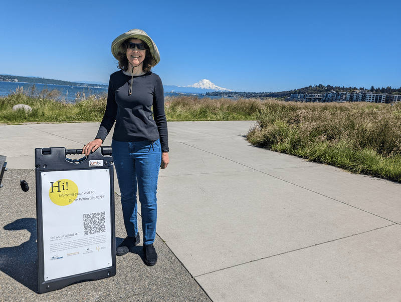 A researcher stands beside a sign for a survey with Mount Rainier in the background.