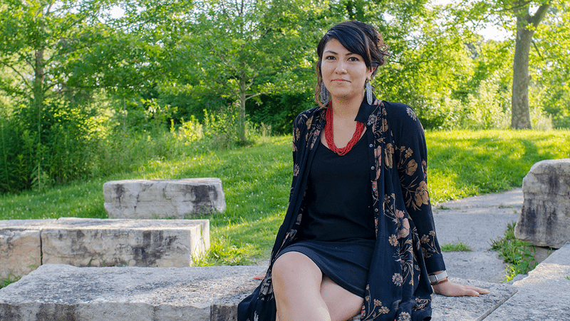L. Irene Compadre sits on a stone bench in front of a walkway, grass and trees.
