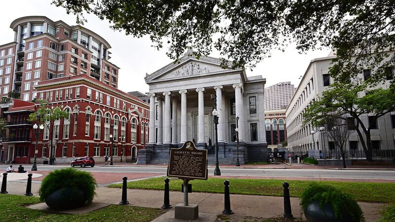 Gallier Hall in New Orleans, featuring a historical marker plaque for Lafayette Square in the foreground. The grand neoclassical building has tall columns and a triangular pediment.