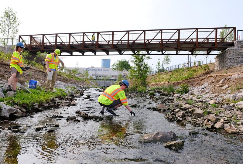 A researcher in a safety vest and hard hat squats in a stream to collect a water sample while two other researchers look on.