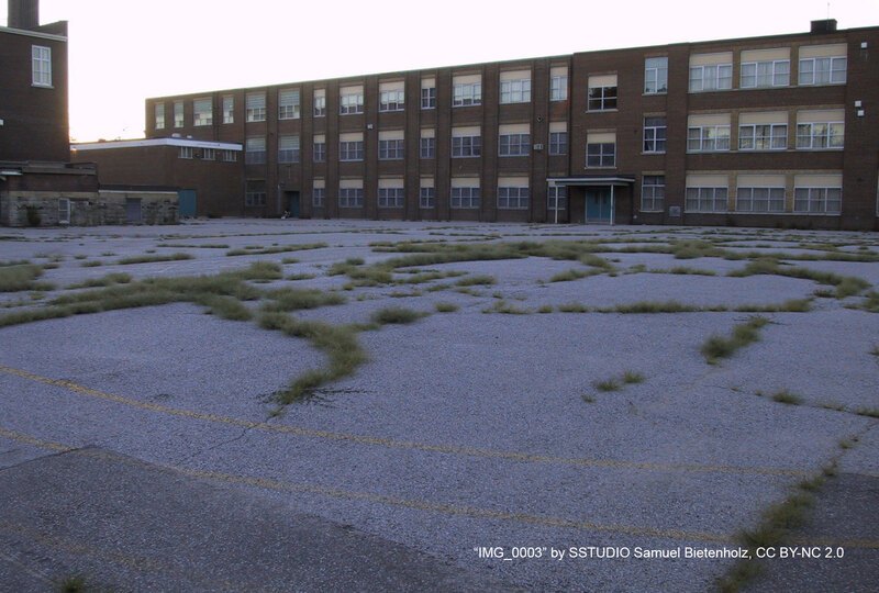 An empty asphalt schoolyard with grass growing through the cracks with the school building visible in the background