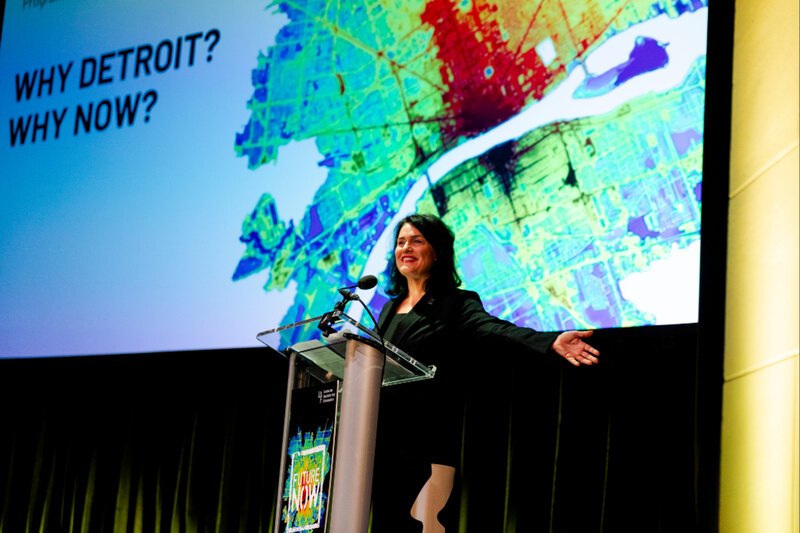 speaker standing at a podium infront of a projected screen with the words "Why Detroit? Why Now?"