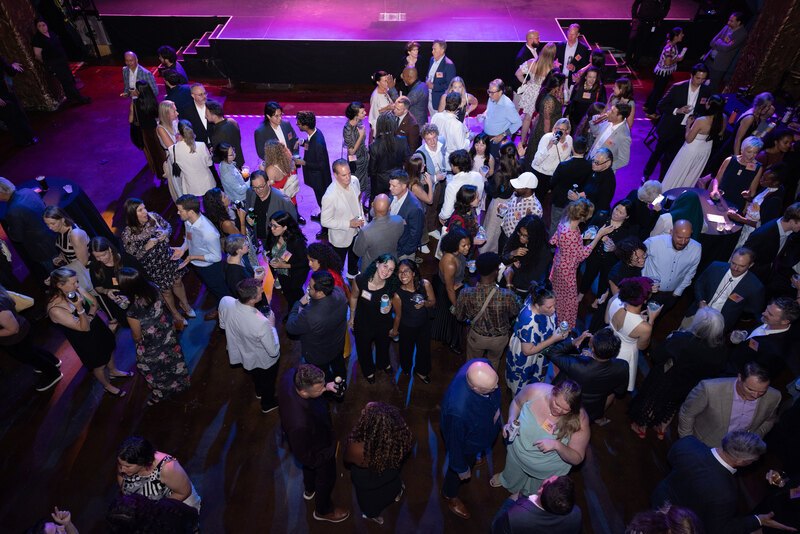 A large crowd mingles at an event inside an ornate theater, illuminated by purple and red lighting