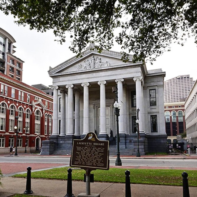Gallier Hall in New Orleans, featuring a historical marker plaque for Lafayette Square in the foreground. The grand neoclassical building has tall columns and a triangular pediment.