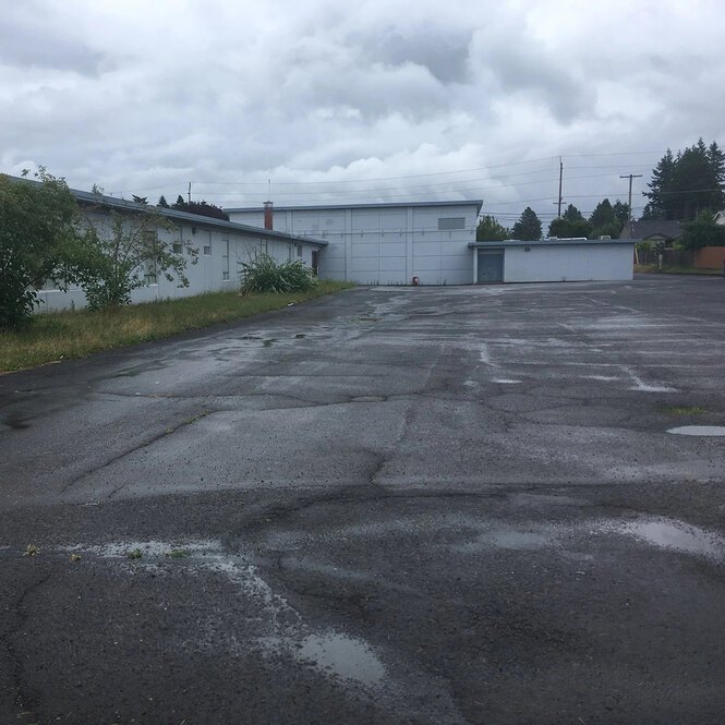 A large, empty asphalt area next to a school building with small puddles and visible cracks in the pavement