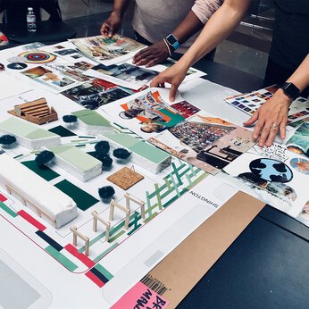 A project model sits on a table while many hands flip through pages of magazines and other colorful paper to add