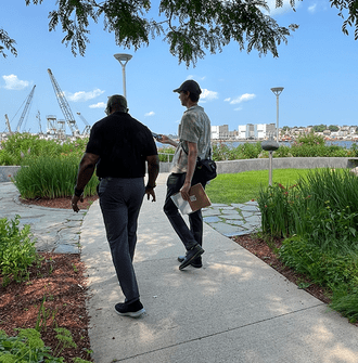 A researcher interviews a plaza user with a river, boats, and loading cranes in the background