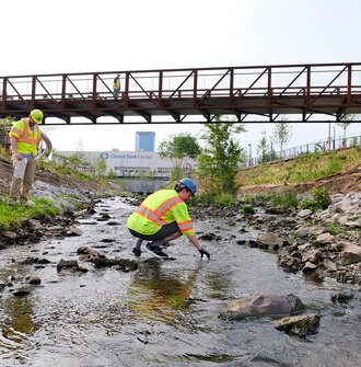 A researcher in a safety vest and hard hat squats in a stream to collect a water sample while two other researchers look on.