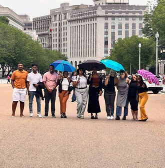 10 LAF Ignite students pose smiling on a busy pedestrian street with some holding umbrellas