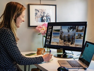 A woman watches a presentation on her large monitor while writing and interacting with coworkers on a laptop.