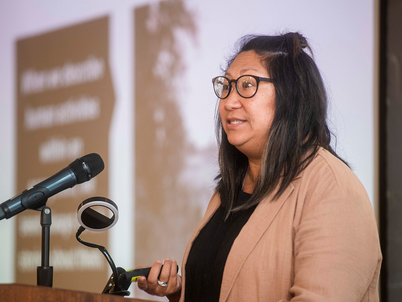 Claire Napawan at a podium with a microphone presenting. A blurred slide image is in the background.