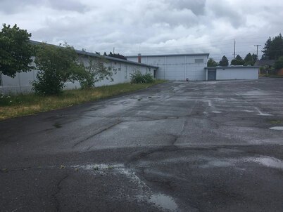 A large, empty asphalt area next to a school building with small puddles and visible cracks in the pavement