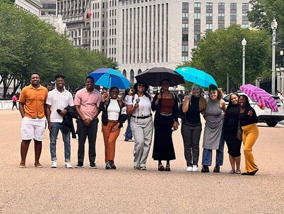 10 LAF Ignite students pose smiling on a busy pedestrian street with some holding umbrellas