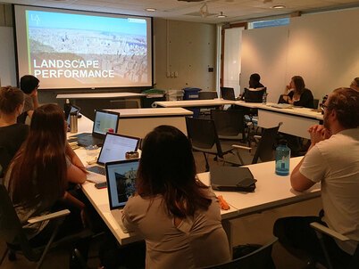 People sit at tables arranged in a U-shape looking at a screen with and image of New York City and the word "Landscape Performance"