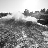 Waves crash over a breakwall near a road and buildings