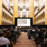 The stage, screen, and panelists in front of a seated audience amidst the columns and arches of the National Building Museum's Great Hall