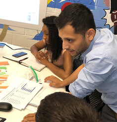 A man in a blue collared shirt leans on a table. A woman sits to his right. The table in front of them has scattered papers with charts and graphs.