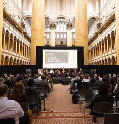 The stage, screen, and panelists in front of a seated audience amidst the columns and arches of the National Building Museum's Great Hall