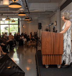 A seated crowd watches Lucinda Sanders on stage behind the podium in front of a screen