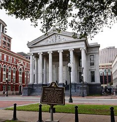 Gallier Hall in New Orleans, featuring a historical marker plaque for Lafayette Square in the foreground. The grand neoclassical building has tall columns and a triangular pediment.
