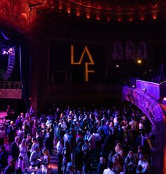 A large crowd mingles at an event inside an ornate theater, illuminated by purple and red lighting, with an illuminated LAF logo projected on the stage backdrop.